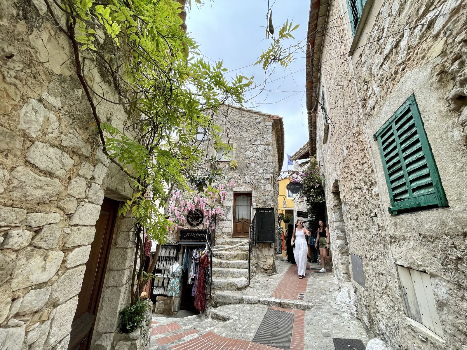 Eze village — narrow stone alley, old shopfronts, Sucheta walking, wisteria above